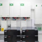 Row of white electrical inverter cabinets with green displays mounted on a wall above several black WECO power units in a clean utility room.