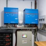 Industrial electrical cabinet room with multiple blue inverters and a gray control panel, overhead cable trays and wiring.