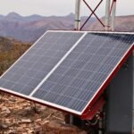 Solar panels mounted on a metal frame in a rocky desert landscape with distant mountains in the background, angled toward the sky.