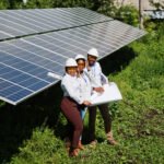 Three workers in hard hats and safety gear stand beside a large solar panel array, reviewing blueprints outdoors.